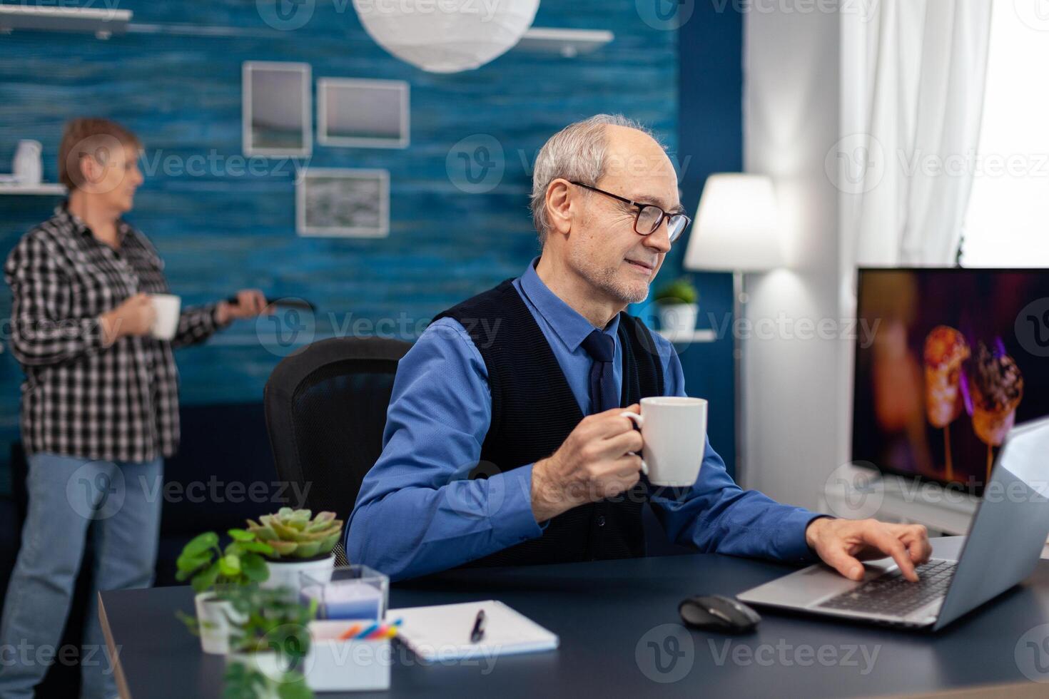 Senior businessman holding cup of coffee working on laptop. Elderly man entrepreneur in home workplace using portable computer sitting at desk while wife is holding tv remote. photo