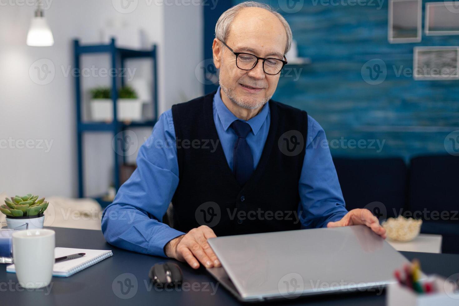 Businessman with gray hair opening laptop to start working. Elderly man entrepreneur in home workplace using portable computer sitting at desk while wife is holding tv remote. photo