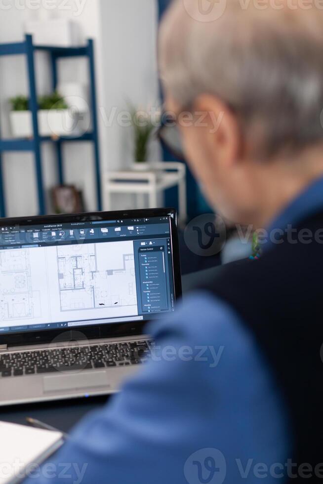 Overshoulder shot of senior architect working on laptop to finish a project. Experienced construction engineer designer looking at house blueprints on laptop computer from home office. photo
