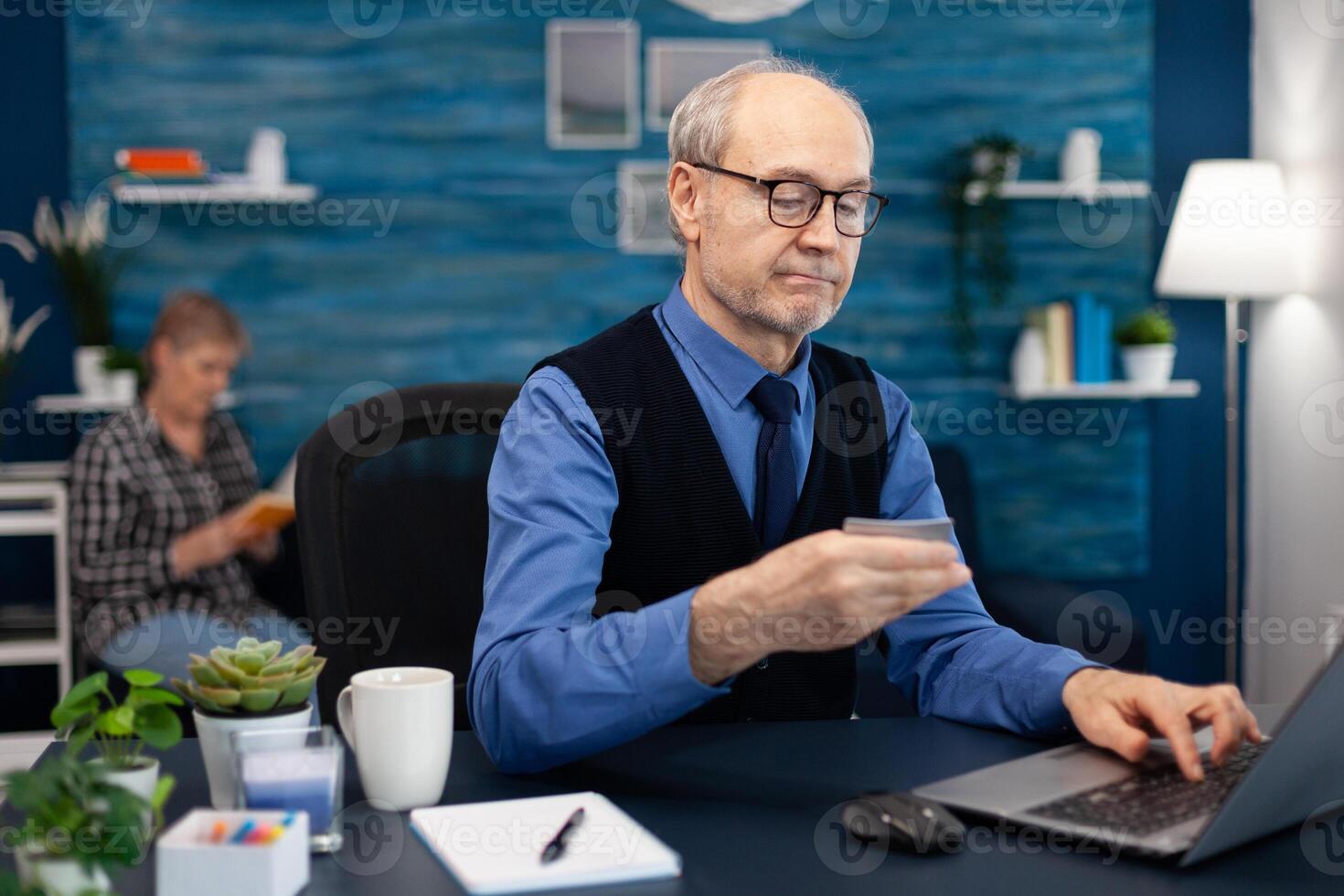 Senior man using credit card to check bank account. Elderly man checking online banking to make shppping payment looking at laptop while wife is reading a book sitting on sofa. photo