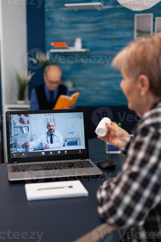 Senior pensioner listening doctor talking about treatment during online consultation. Elderly woman discussing with healthcare practitioner in the course of remote call and husband is reading a book on sofa. photo