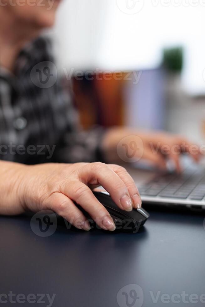 Close up of old entrepreneur lady using mouse in home office. Elderly woman in home living room using moder technoloy laptop for communication sitting at desk indoors. photo
