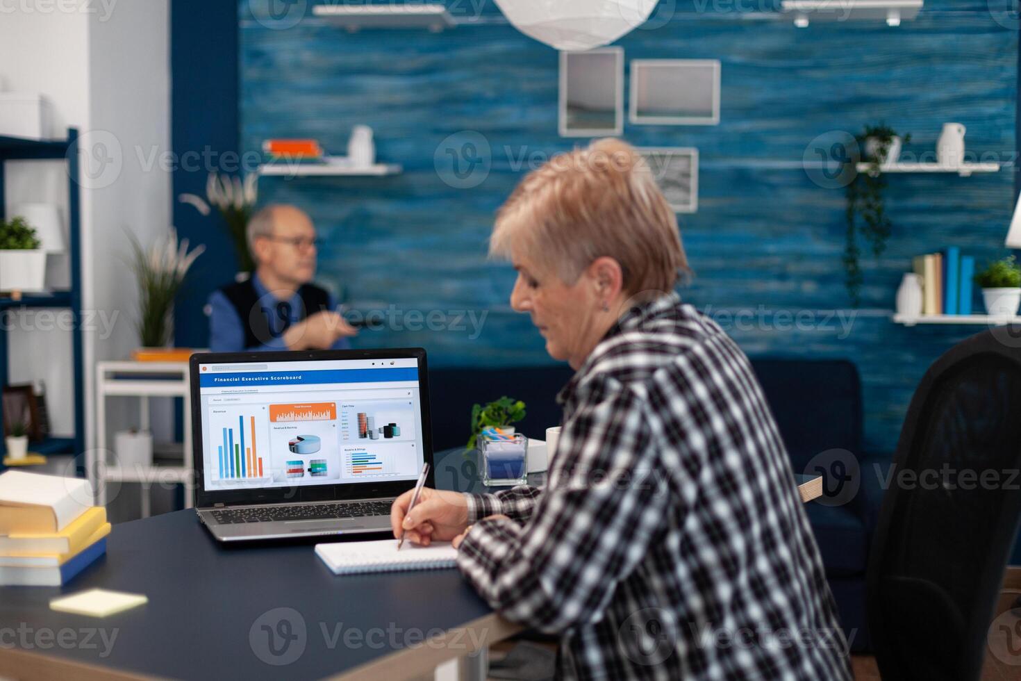 Elderly senior woman taking notes in notebook while working on laptop in cozy living room. Adult internet finance computer caucasian mature technology glasses browsing connection retored retirement photo