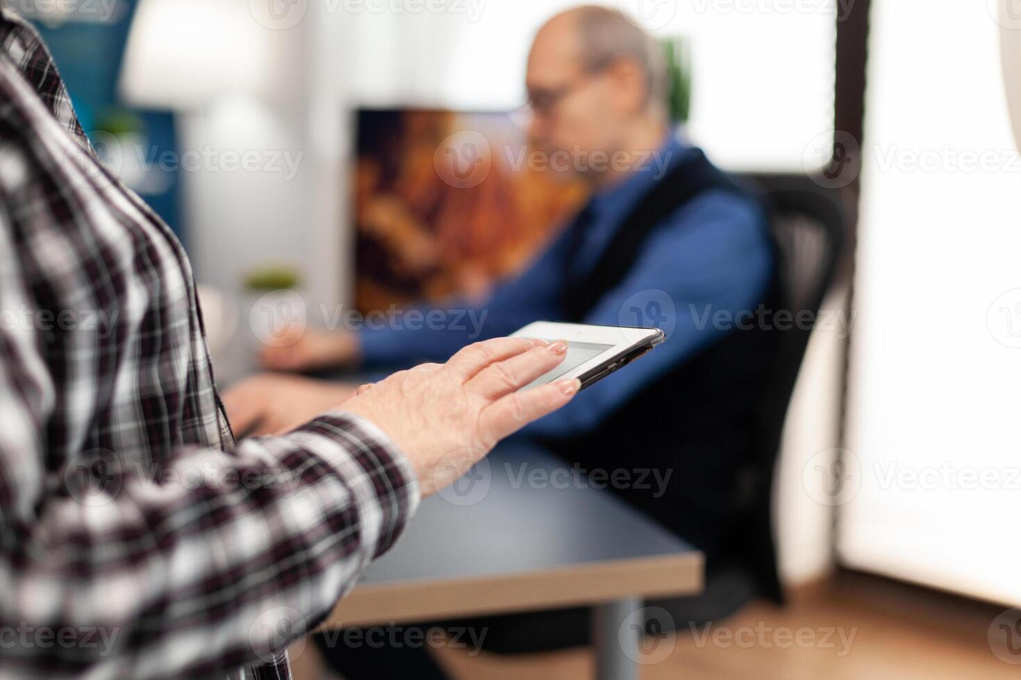 Close up of senior woman hand scrolling on digital device in home. Close up of elderly woman using moder technoloy tablet pc in home living room and husband working on laptop computer. photo