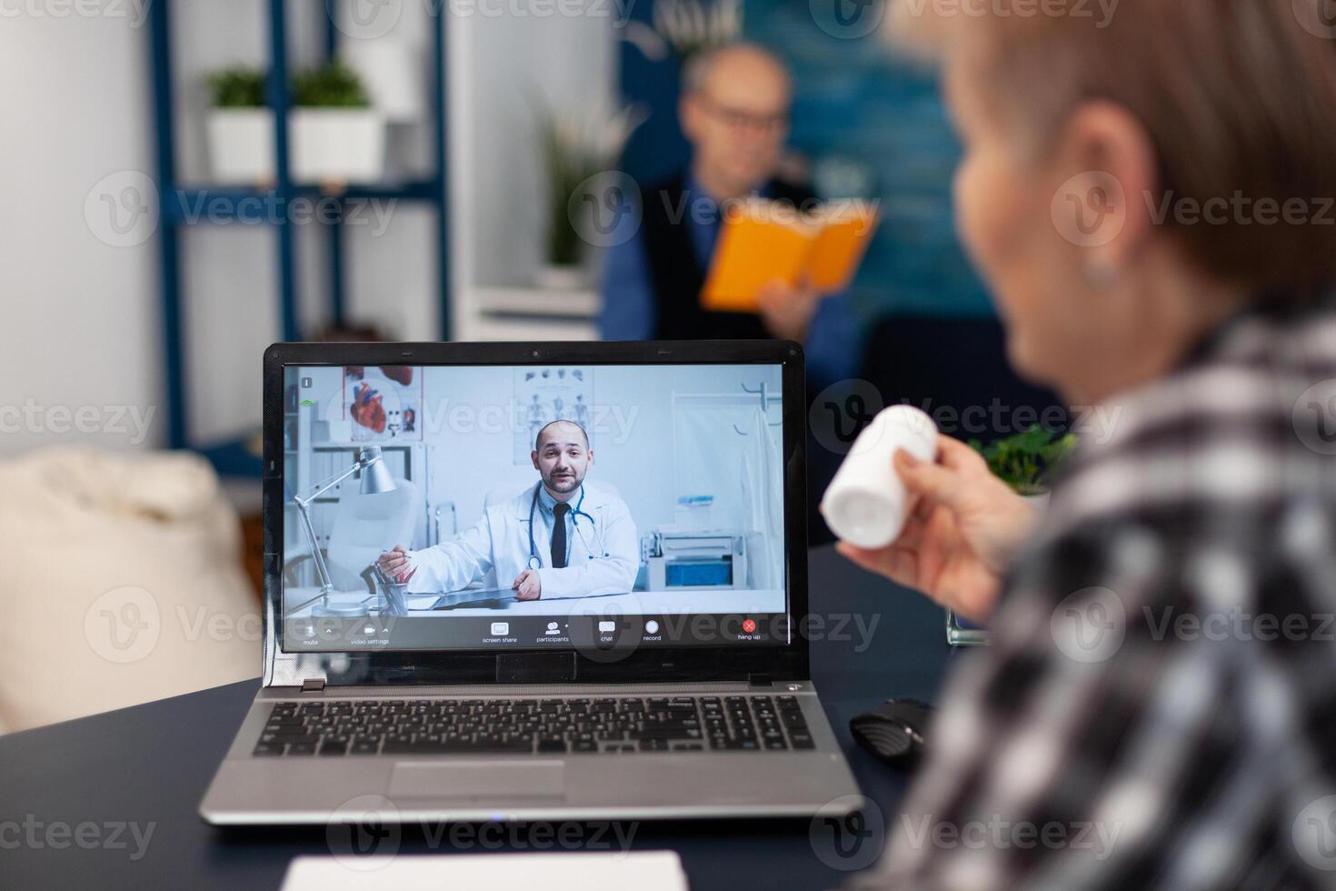 Older woman listening doctor indication about health treatment during call. Elderly woman discussing with healthcare practitioner in the course of remote call and husband is reading a book on sofa. photo