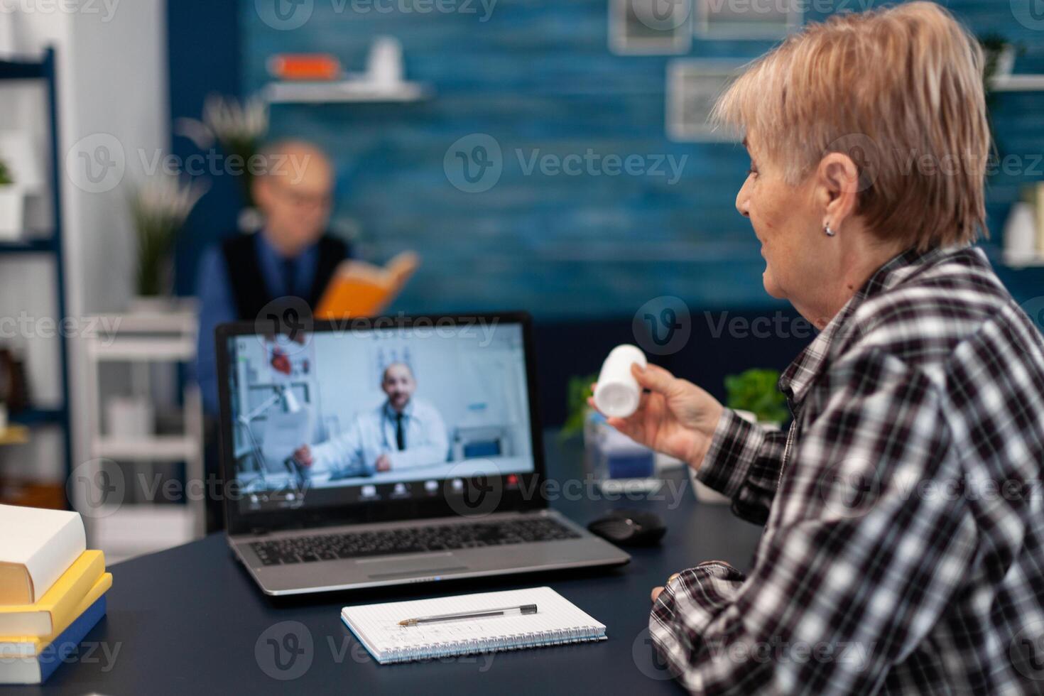 Senior woman showing pills bottle during medical online conference. Elderly woman discussing with healthcare practitioner in the course of remote call and husband is reading a book on sofa. photo