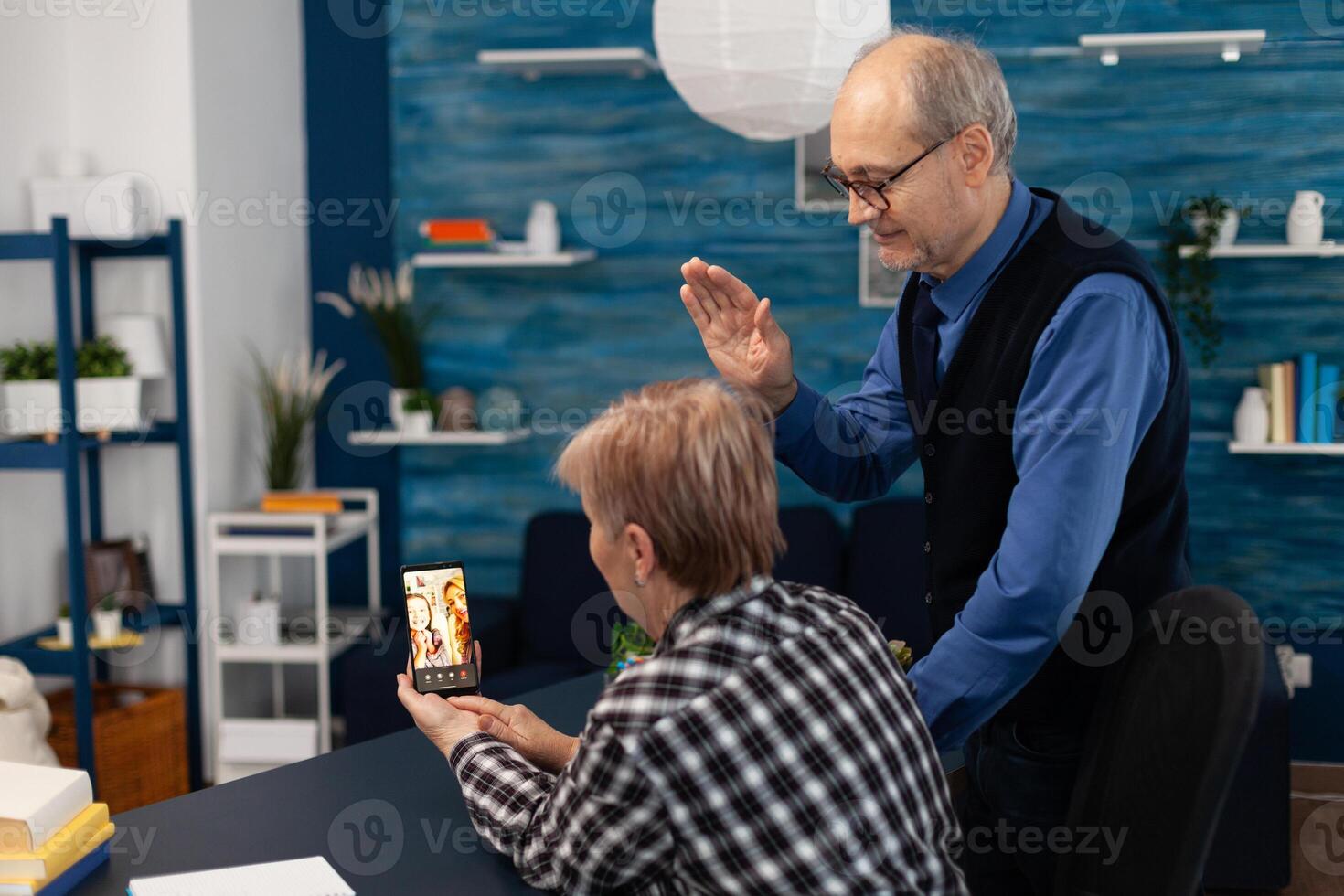 Senior man waving at smartphone camera during call with relatives. Happy grandparents communicating with family via online web internet conference using modern internet technology while sitting in the living room photo