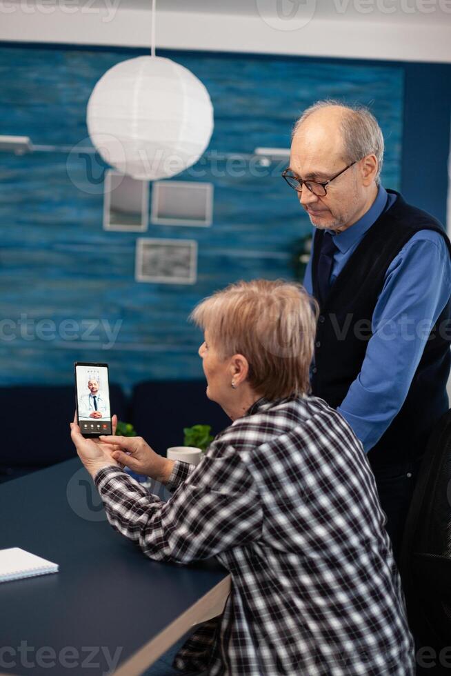 Senior sick man and woman talking with doctor during call. Elderly woman and woman discussing with healthcare practitioner in the course of remote call and husband is reading a book on sofa. photo