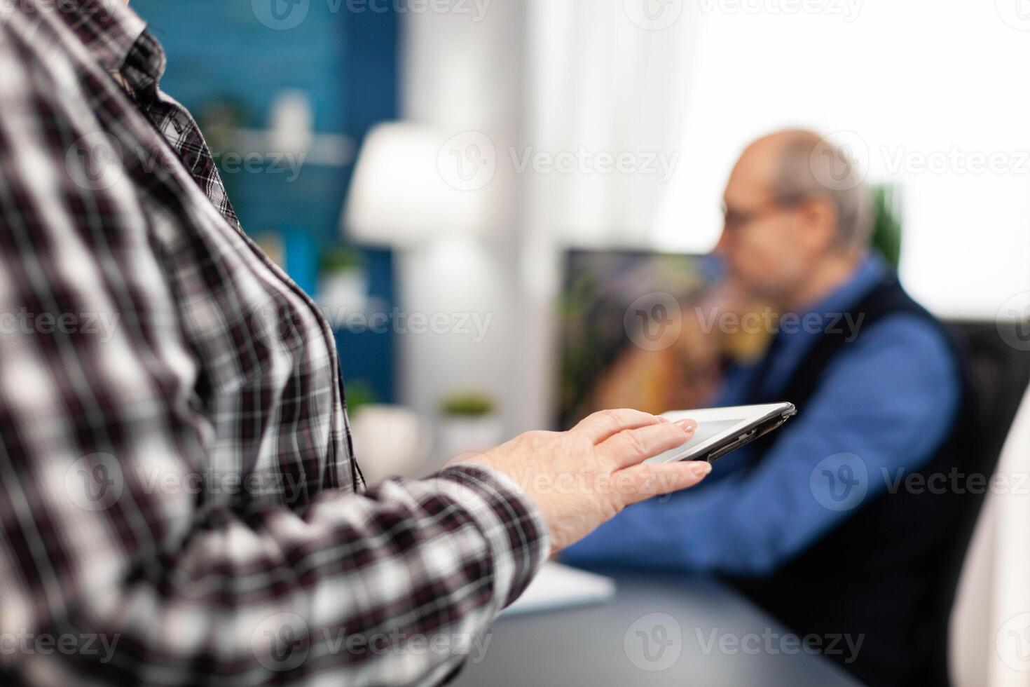 Close up of senior woman hand scrolling on digital device in home. Close up of elderly woman using moder technoloy tablet pc in home living room and husband working on laptop computer. photo