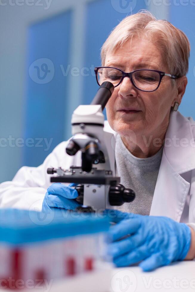Senior chemist researcher in white coat looking in high end microscope for disease expertise. Modern laboratory for scientific research with professional equipment for virus study and diagnosis biotechnolog development photo