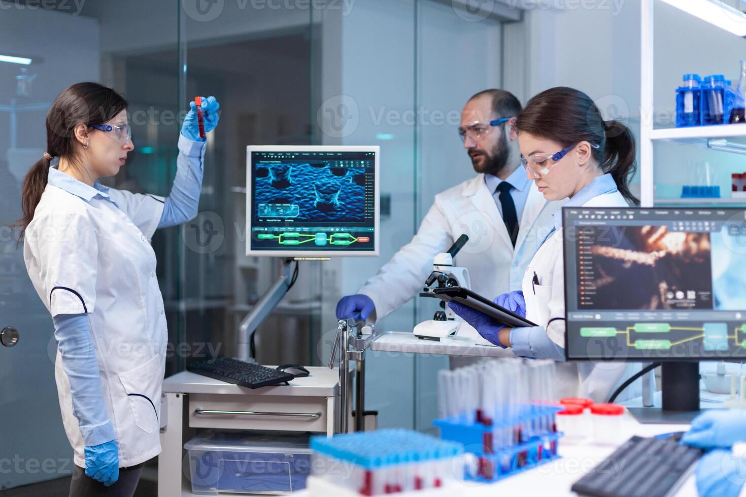 Team of medical research scientists conducting vaccine development with help of high tech, test tubes, micropipette and writing down analysis results on computer in modern equipped laboratory at night photo