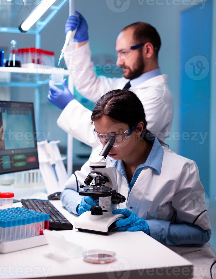 Scientist looking at blood sample through microscop and assitant taking liquid with micropipette. Modern lab for scientific research with professional equipment for vaccine development diagnosing virus evolution. photo