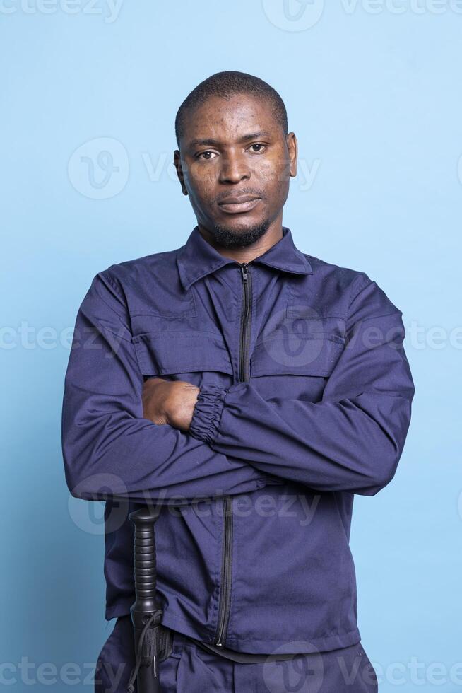 Portrait of serious proud security guard posing in uniform on camera, standing with arms crossed against blue background. African american armed bodyguard works on ensuring protection. photo