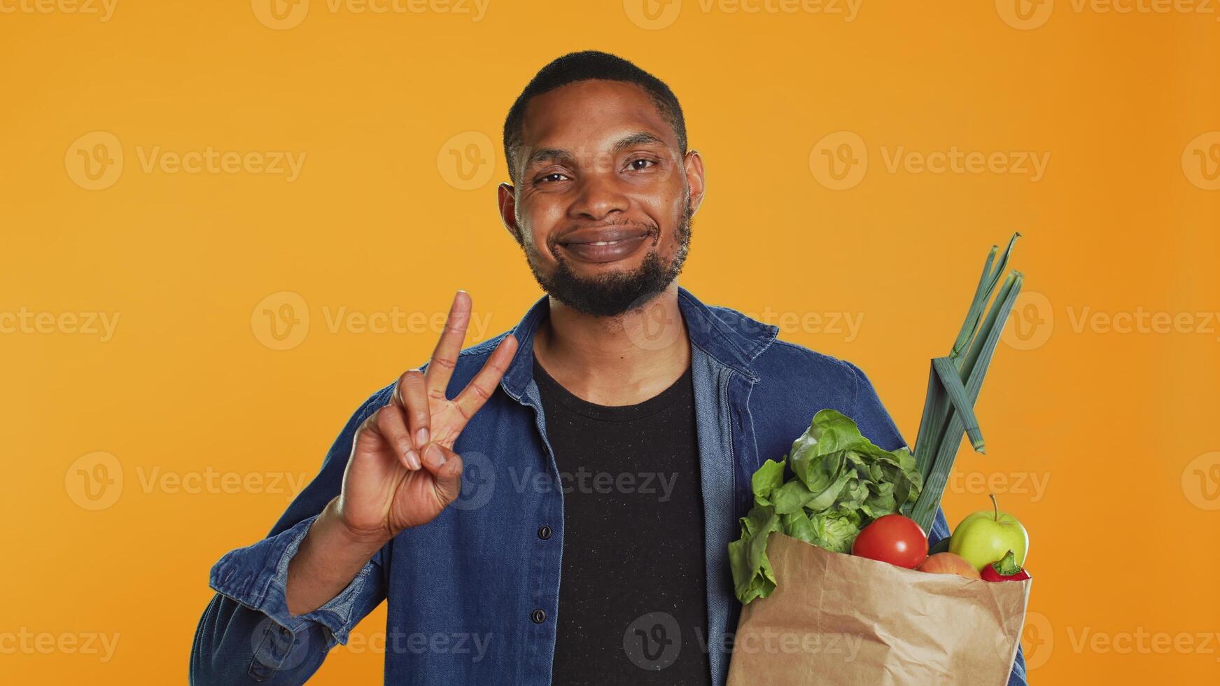 Young adult showing peace sign gesture and holding organic groceries, recommending sustainable lifestyle and homegrown produce. Peaceful relaxed guy carries eco friendly goods. Camera A. photo
