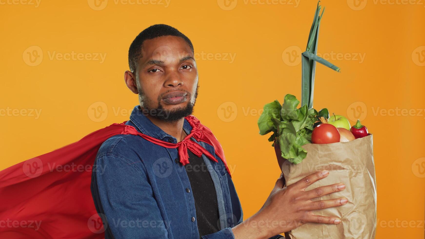 Young adult acting as a superhero with a red cape presenting homegrown produce in a paper bag, advocating for sustainable lifestyle concept. Person supporting organic food. Camera A. photo