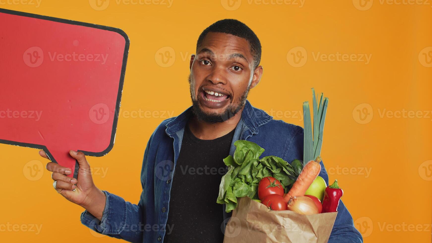 Young man showing a speech bubble empty cardboard sign to create an ad for sustainable lifestyle healthy eating. Guy doing a promotional advertisement for bio ripe fruits and veggies. Camera A. photo