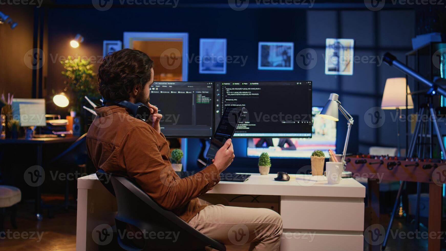 Software developer programming in home office, crosschecking lines of code between tablet and PC screen. IT admin holding portable device, using it as reference while coding on computer, camera B photo