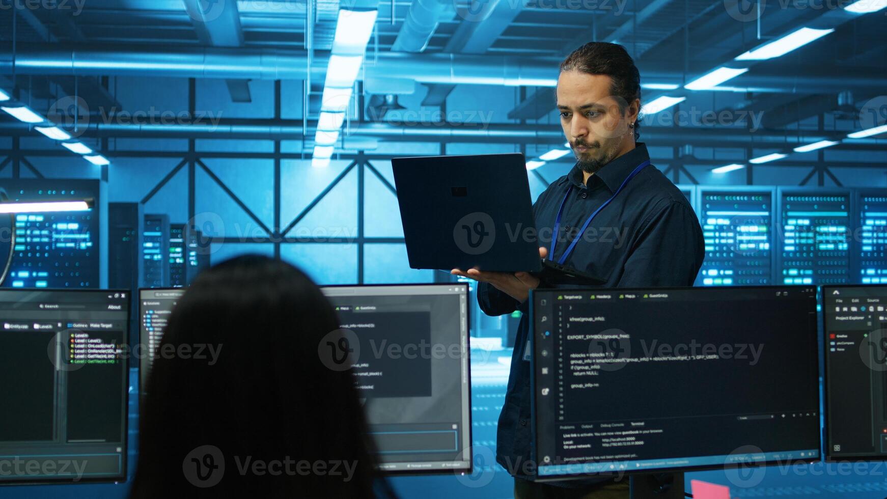 Engineer typing on laptop in server hub, checking recovery plan, monitoring data center energy consumption across components. Technician inspecting rackmounts, doing routine disk checks photo