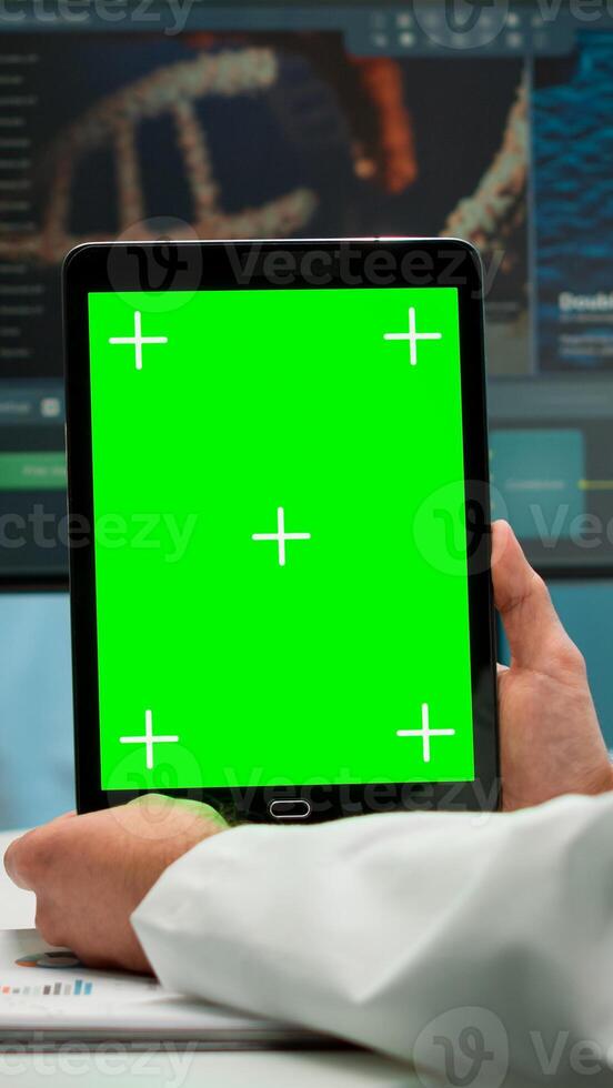 Close up of biochemist sitting at workplace in laboratory using green mock-up screen tablet with chroma key display while coworker working in background bringing blood sample. photo
