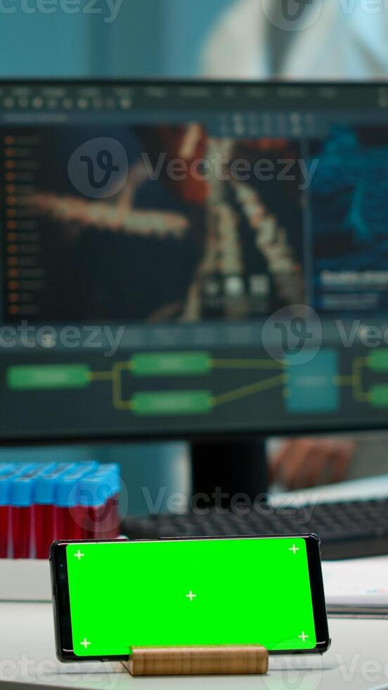 Microbiologist bringing blood samples in modern lab putting them near smartphone working with green chroma key screen placed on desk. Team of biotechnology scientists developing drugs in background photo