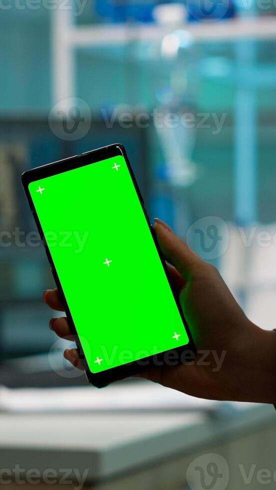 Close up of biochemist holding and looking at phone with chroma key display standing in biological lab while nurse in white coat bringing blood sample. Scientist using phone with mockup, green screen photo
