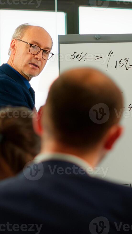 Elderly businessman giving presentation to multiethnic team of colleagues using flip chart, drawing, explaning financial graphs in meeting room. Manager interacting with audience at corporate workshop photo