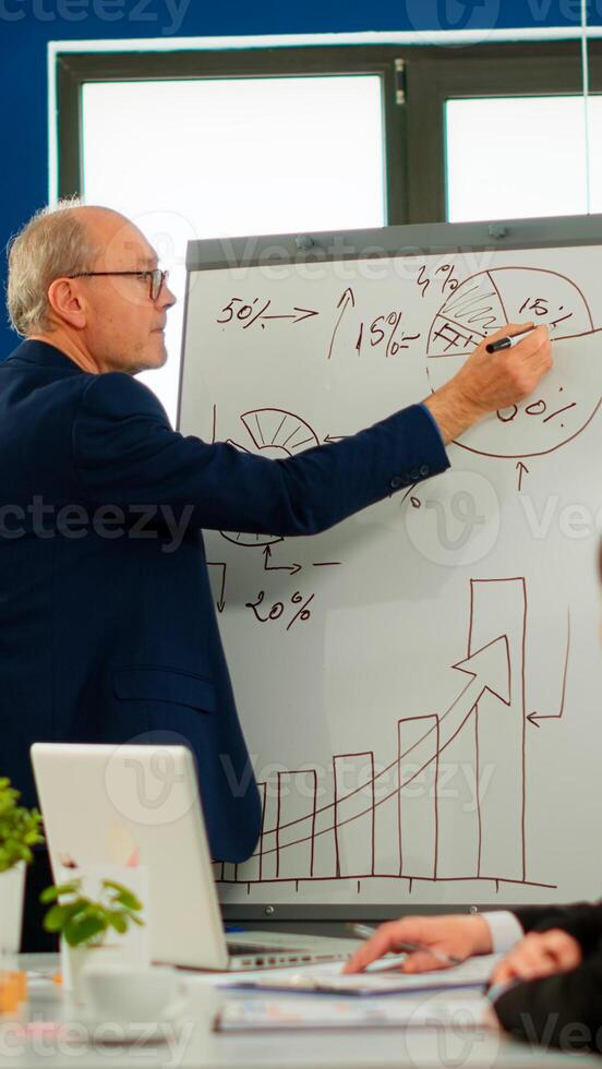 Senior businessman in eyeglasses and formal suit drawing financial plan on flipchart and explaining to interracial team of colleagues while giving presentation during conference meeting, brainstorming photo
