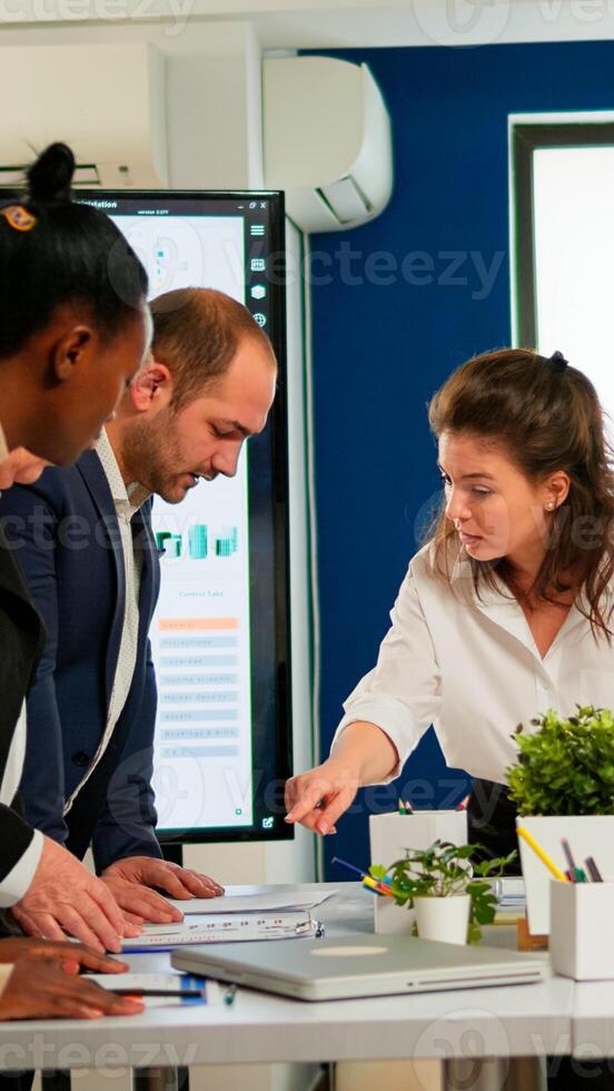 Diverse team of workers talking, looking at documents and analyzing graphs data standing at conference desk in start up company. Professional colleagues team discussing marketing project brainstorming photo