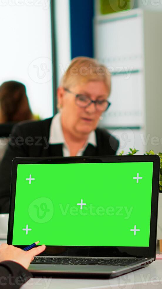 Over the sholder of manager sitting at meeting desk looking at laptop with green screen mockup while diverse team working on background. Multiethnic people planning project on chroma key display photo