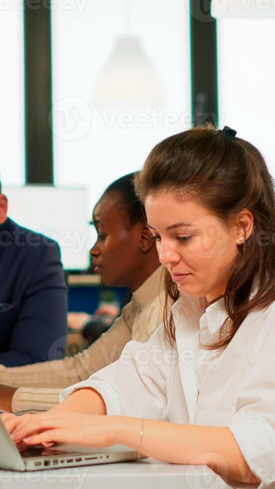 Satisfied business woman answering emails typing on laptop smiling sitting at desk in busy start up office while diverse team analyzes statistics data. Multiethnic team working at new project photo