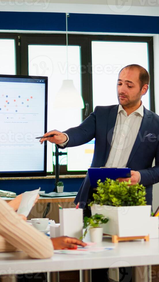 Manager pointing on digital interactive whiteboard with growth analysis, charts, statistics and data. Diverse people working in broadroom during conference meeting discussing company problems. photo
