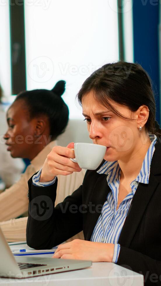 Concentrated manager typing on laptop sitting at desk in start up business office drinking coffee while diverse colleagues working in background. Multiethnic coworkers planning new financial project. photo