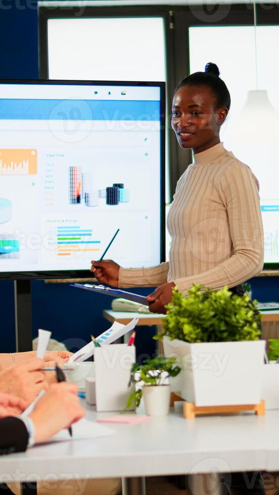 Black woman worker explaning company statistics in front of colleagues, briefing group of employees. Multiethnical businesspeople working in professional startup financial company during conference photo