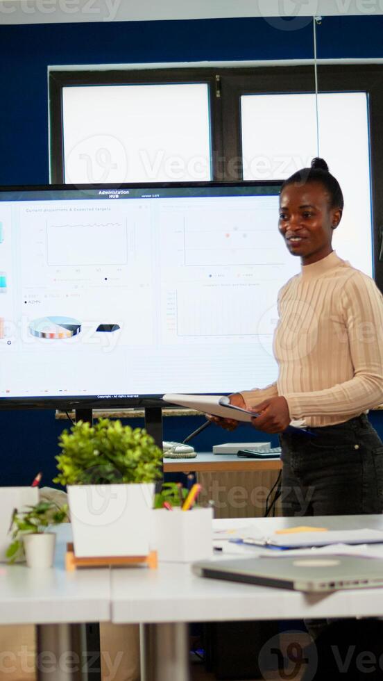 Black analyst using digital interactive whiteboard for presentation to a board of executives lawyers, in corporate meeting room, investors congratulating her clapping. Screen shows company growth data photo