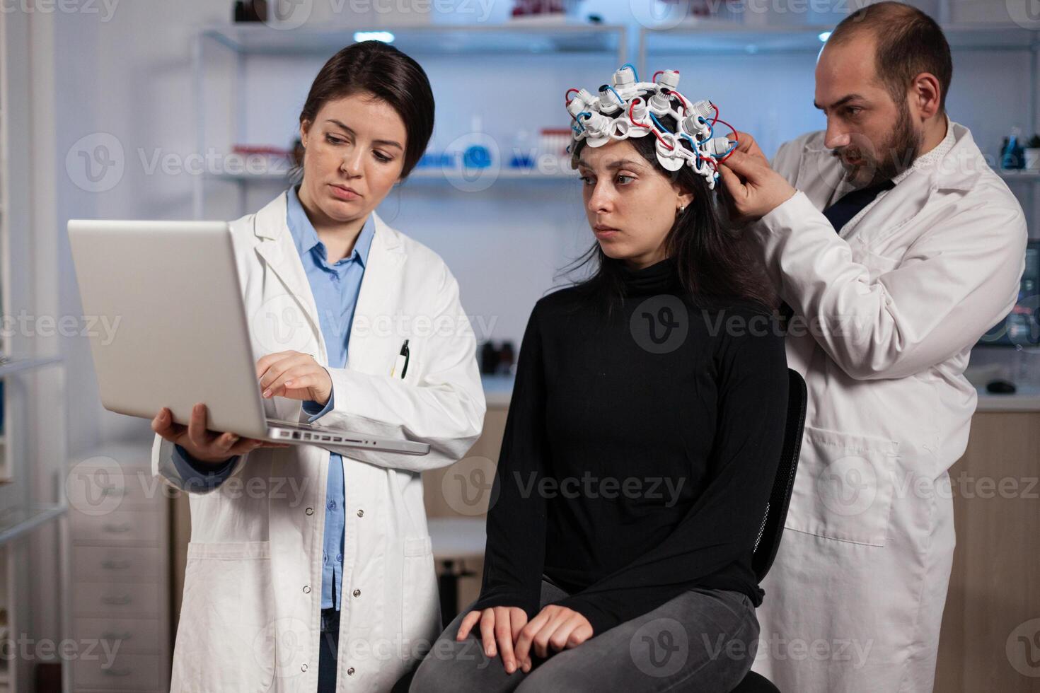 Specialist doctor holding laptop computer showing tomography to woman patient while neurologist man adjusting eeg scanner during neurology experiment. Team of researchers analyzing brain activity photo