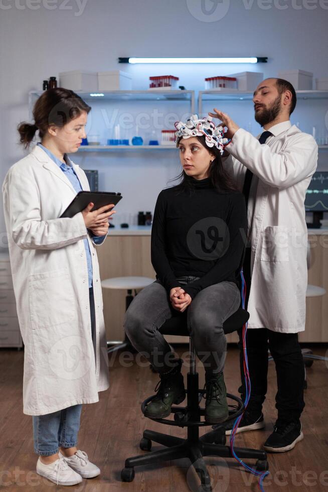 Scientist doctor explaining neurology disease symptoms to woman patient while neurologist researcher adjusting eeg headset during neuroscience experiment in lab. Specialist analyzing brain activity photo