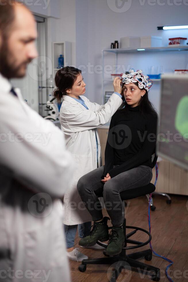 Neurologist doctor analyzing nervous system activity on monitor while specialist doctor adjusting eeg scanner of woman patient during neurology experiment. Medical team monitoring brain evolution photo