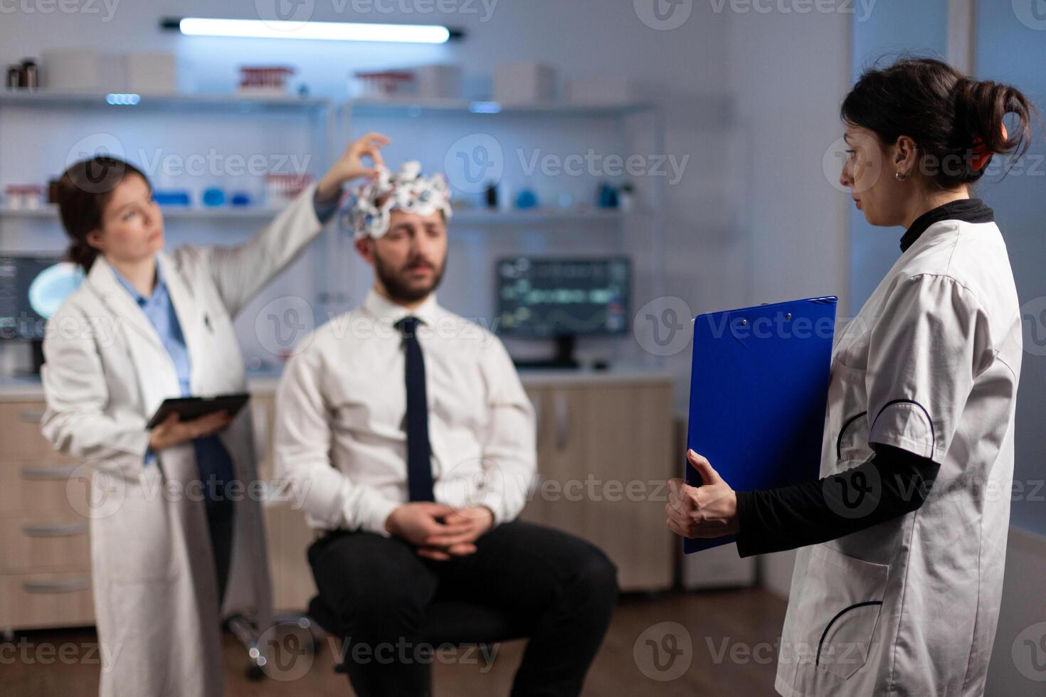 Researcher woman holding clipboard with neuroscience information on it while neurologist doctor adjusting eeg headset monitoring brain activity of man patient. Specialist analyzing nervous system photo