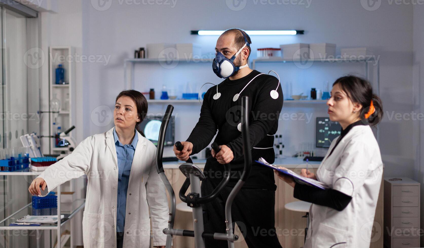 Medical team monitoring body endurance of sportman analyzing EKG data during medicine workout in modern laboratory. Athlete man with electrodes sensor on it running on fitness trainer photo