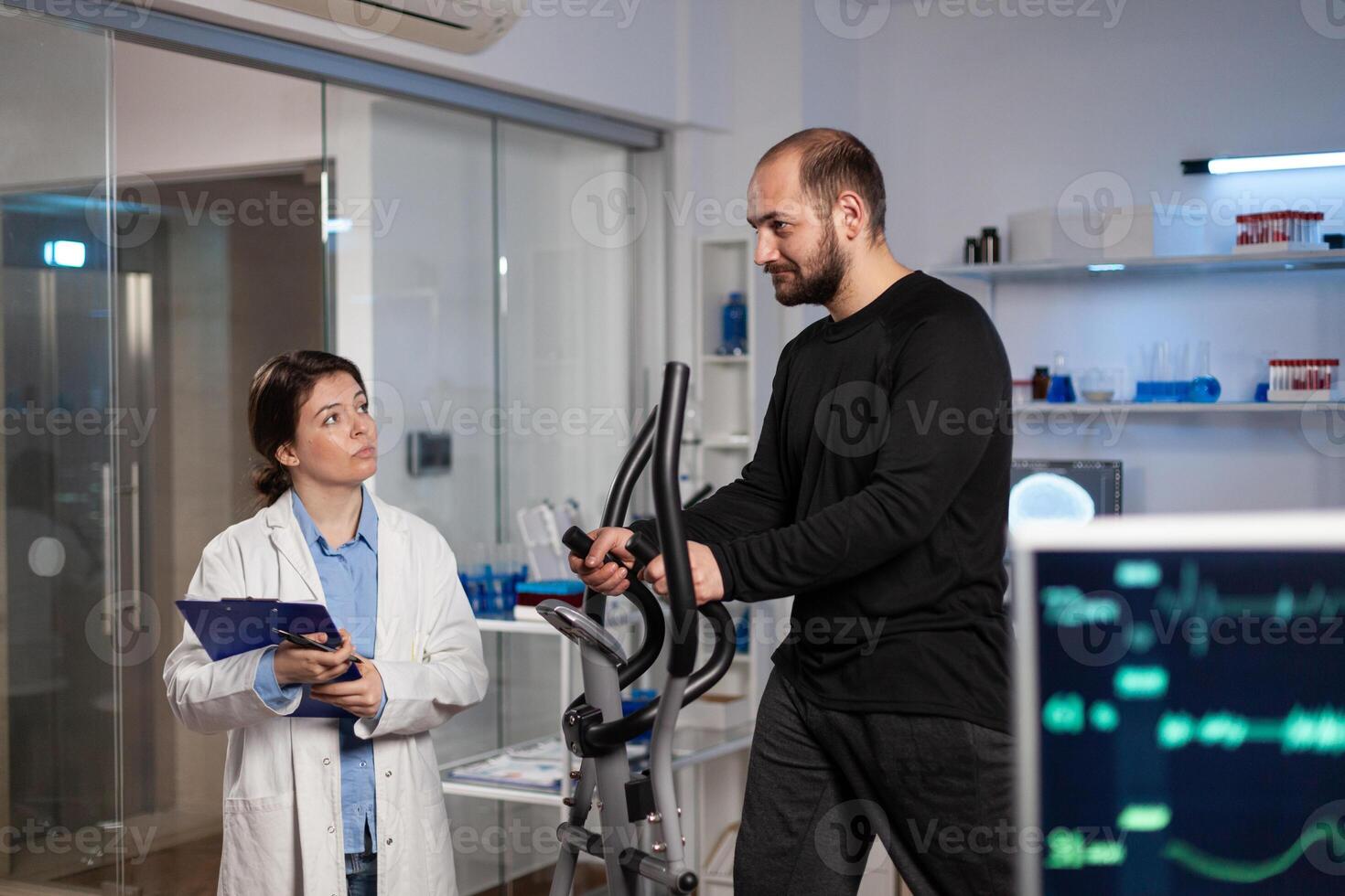 Runner man in sport equipment working at body endurance during medical examination in modern laboratory while woman researcher analyzing EKG data measuring heart rate pulse. Medicine service photo