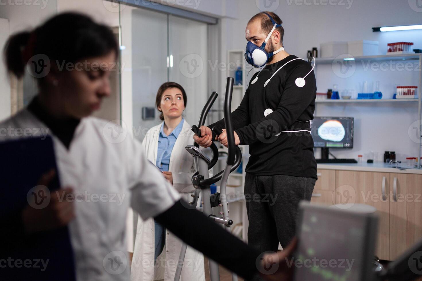 Specialist doctor looking at monitor analyzing heart rate pulse while athlete man running on gym bycle working at body endurance during medical workout in modern lab. Physician monitoring EKG data photo