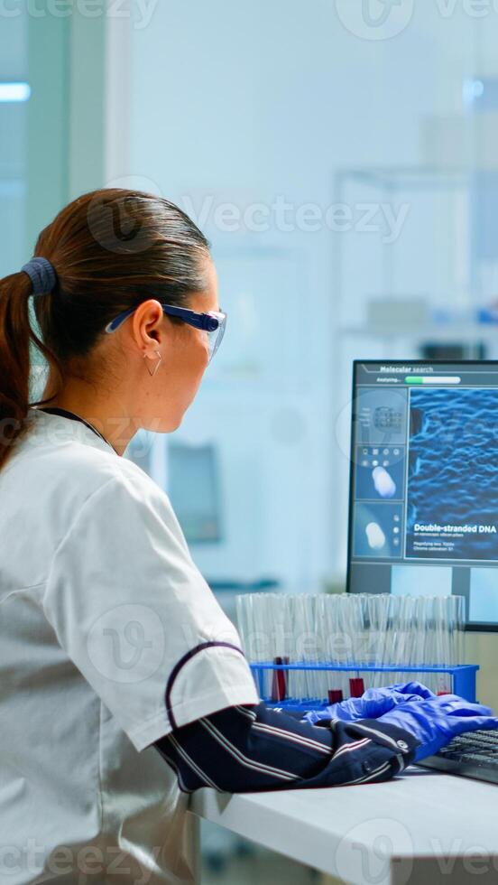 Chemistry doctor analysing blood sample and typing on computer while coworker opening door using digital computer with touch scan. Woman examining virus evolution using high tech and chemistry tools photo