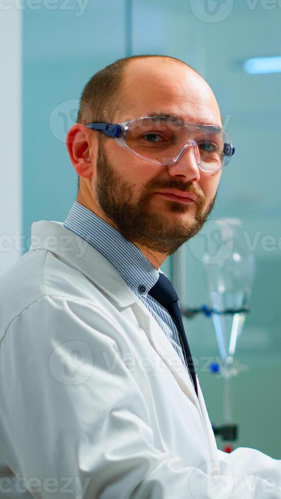 Portrait of scientist man looking at camera sitting in modern equipped laboratory. Scientist doctor examining virus evolution using high tech typing on computer chemistry tools for scientific research photo