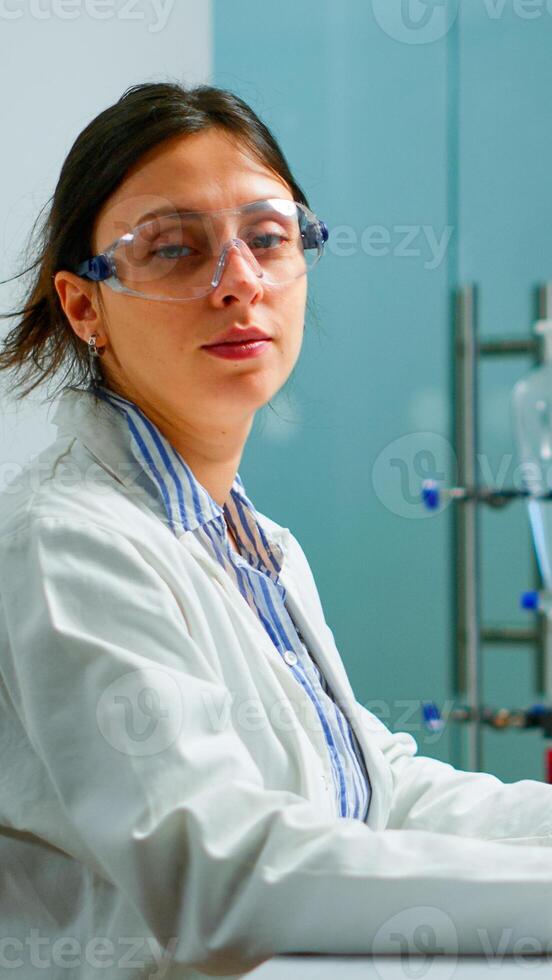 Portrait of tired scientist trying to smile at camera sitting in modern equipped laboratory typing on computer. Chemist examining virus evolution using high tech for scientific research, vaccine photo