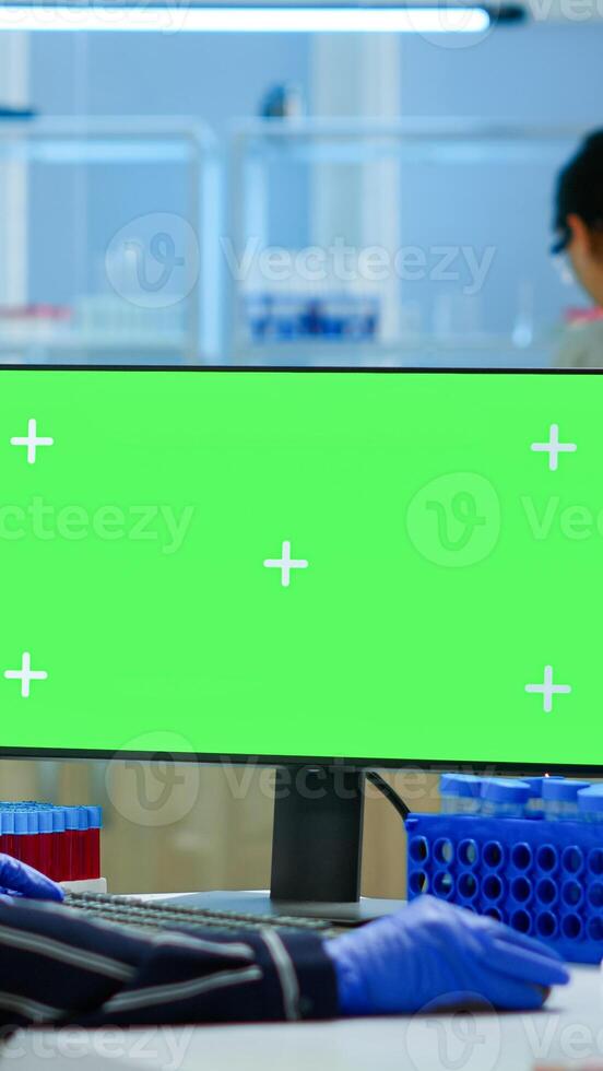 Woman researcher typing at computer with green screen mockup display, template in applied science laboratory. Engineer conducting experiments in background, examining vaccine evolution using high tech photo