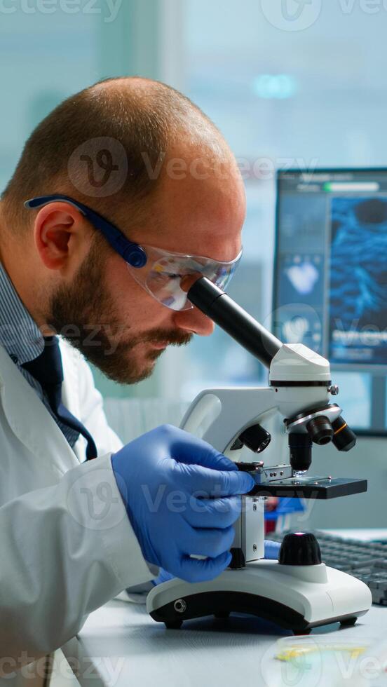 Male scientist looking under microscope in medical development laboratory, entering data in computer. Specialists working on medicine, biotechnology research in advanced pharma lab photo