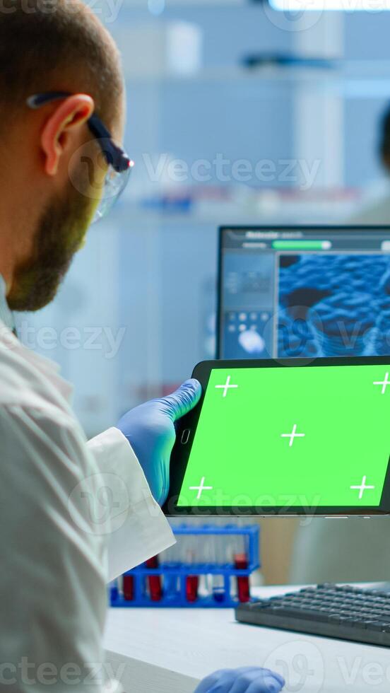 Researcher holding and looking at tablet with chroma key display in modern equipped lab. Team of microbiologists doing vaccine research writing on device with green screen, isolated, mockup display. photo