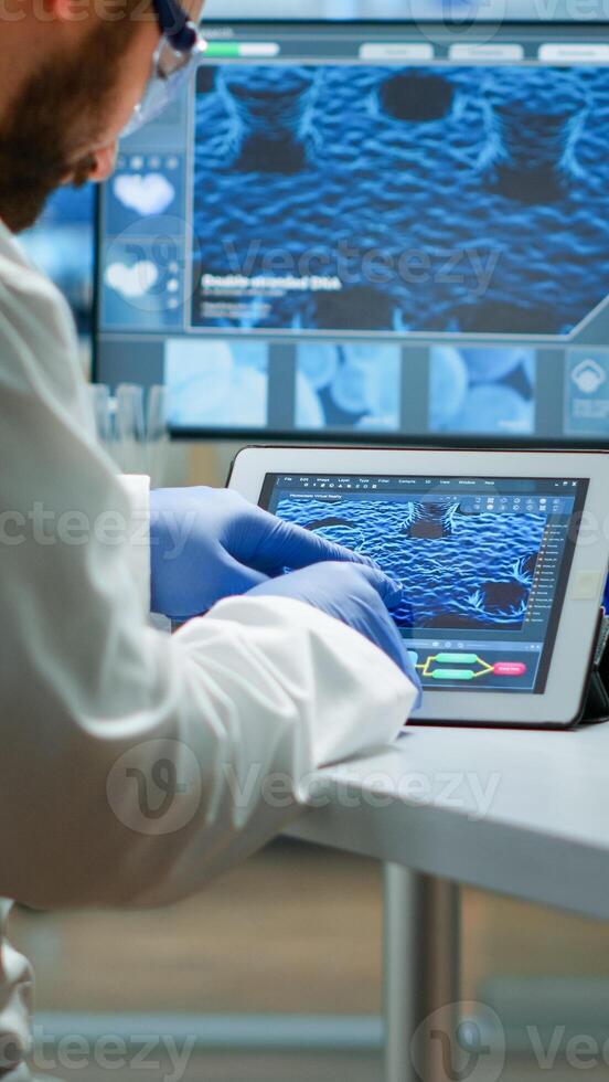 Biochemist checking manifestations of virus looking in tablet with special software in chemistry laboratory. Group of doctors examining vaccine evolution using high tech researching diagnosis photo