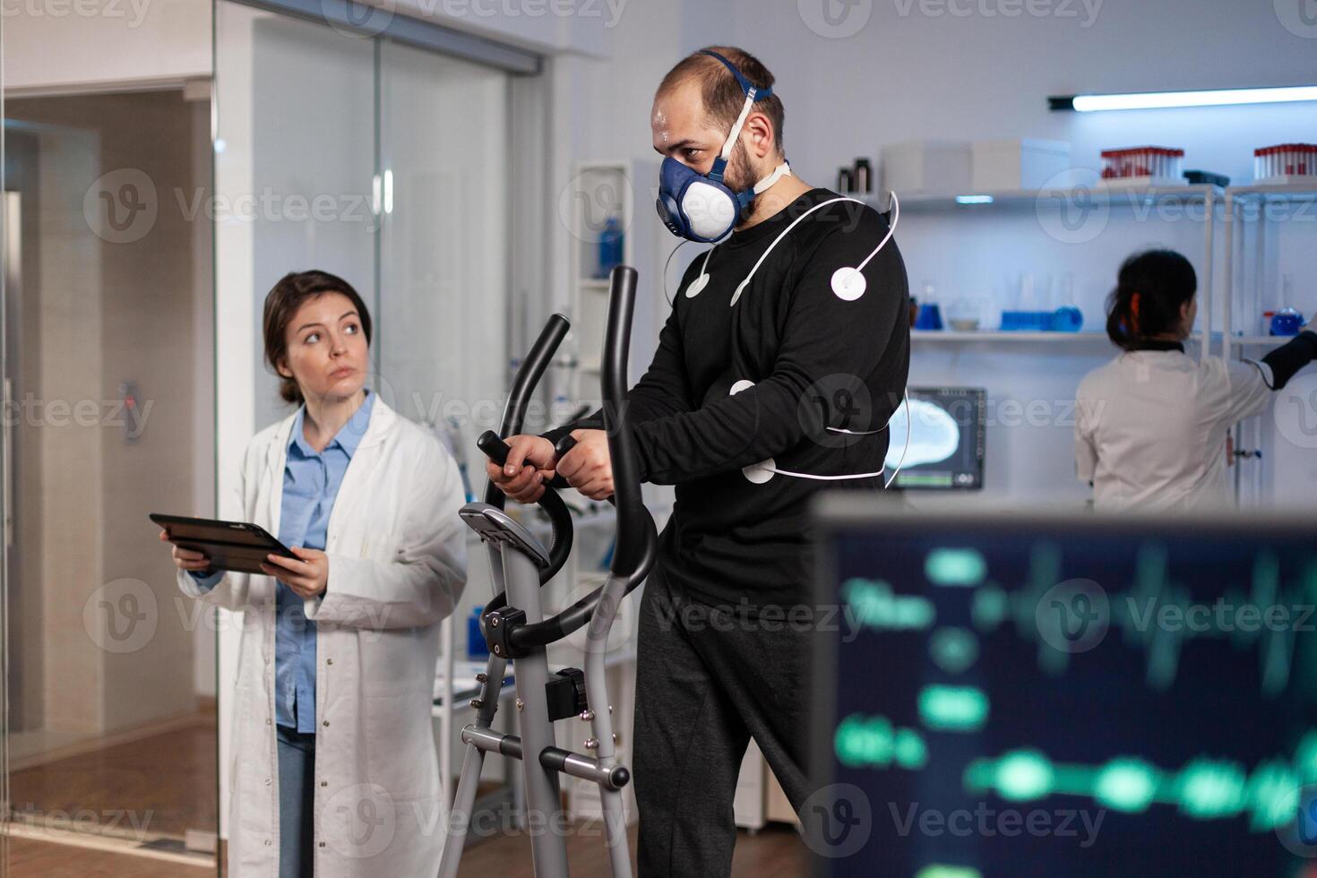 Athlete man with medical electrodes on it running on fitness bycle working at body endurance during medical training in modern laboratory while doctor monitoring EGK data. Heart rate pulse on monitor photo