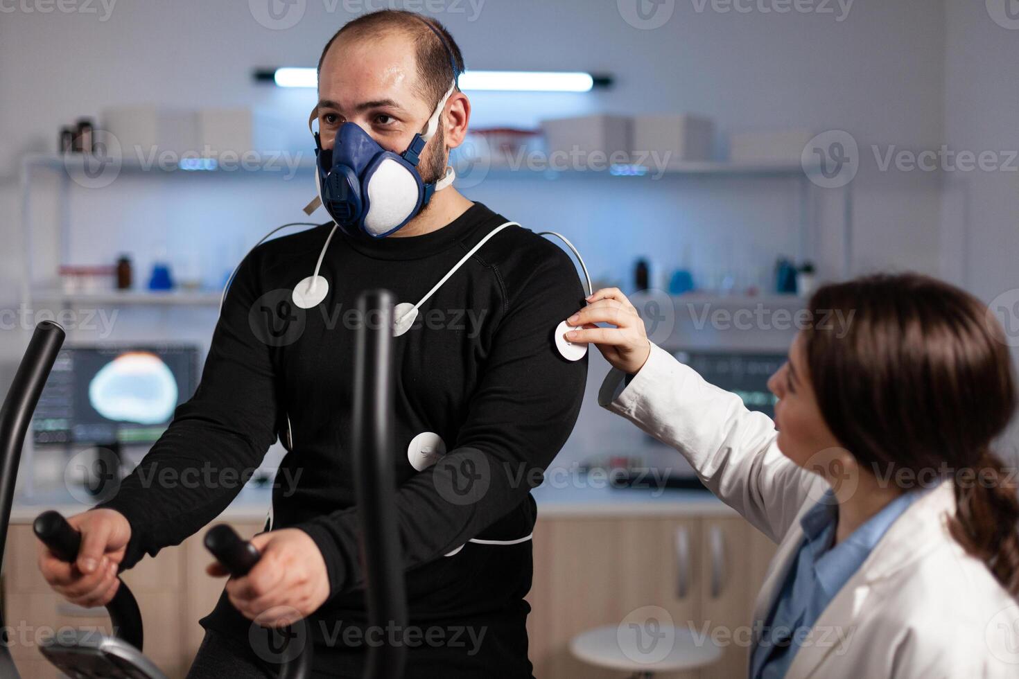 Specialist woman researcher putting electrodes sensor on sportman with mask monitoring EGK data in medical mordern laboratory. Athlete man running on fintess trainer working at body endurance photo